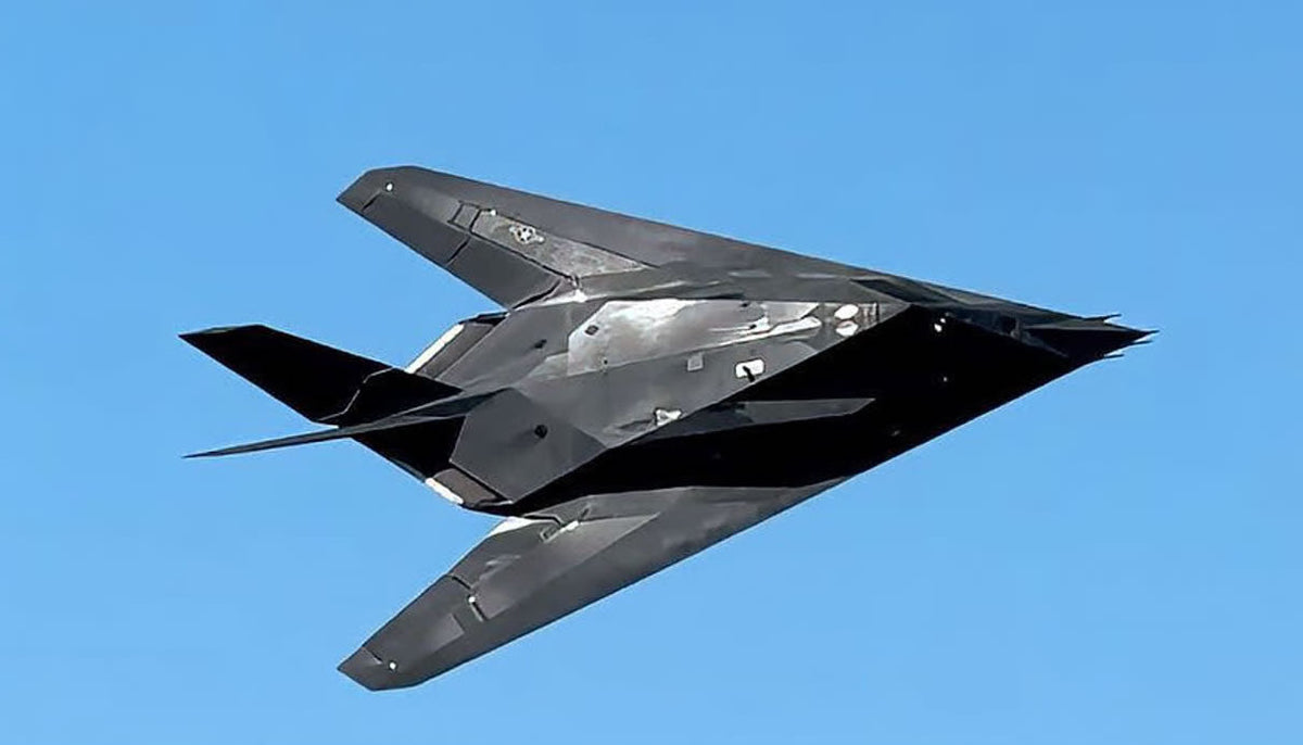 The black faceted 1/72 F-117 Nighthawk diecast model on its display stand on a walnut desk beside its open presentation box, under warm lamp light