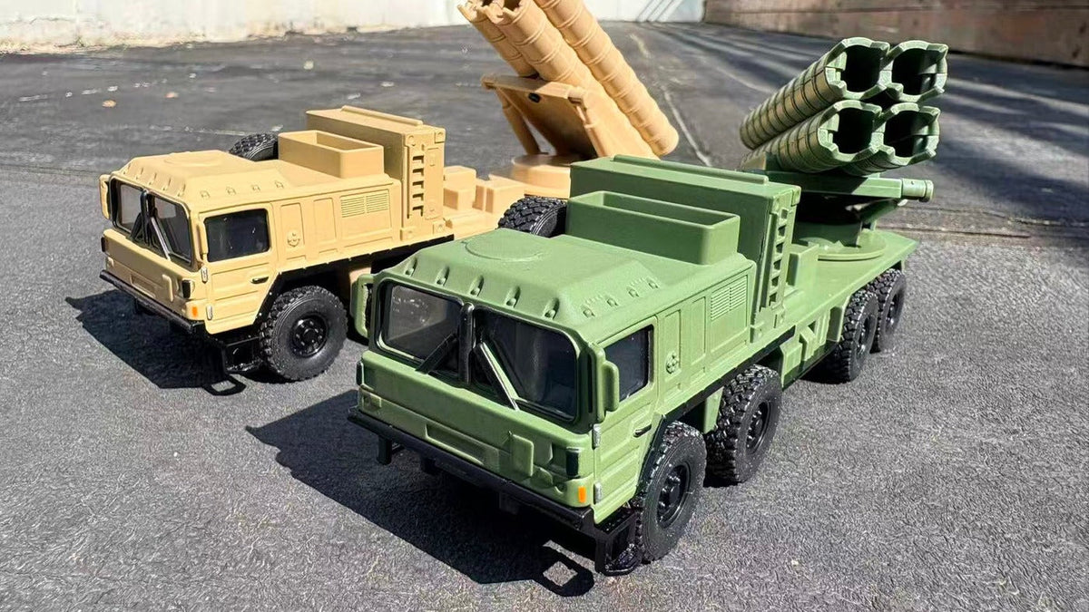 Desert yellow and green 1:64 MAN SX 8x8 diecast tactical truck models side by side on sunlit asphalt, launcher tubes raised
