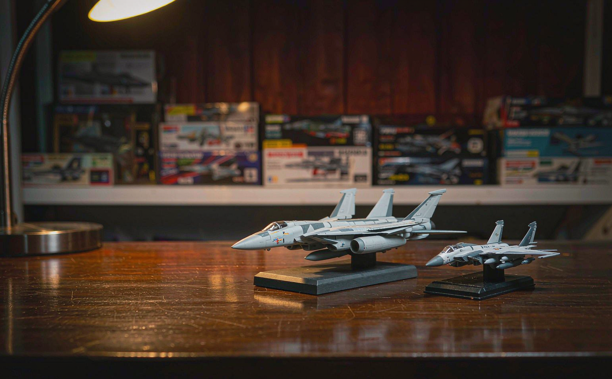 A 1/72 scale diecast F-14 Tomcat and a smaller 1/100 scale diecast fighter model side by side on a wooden desk under warm lamp light